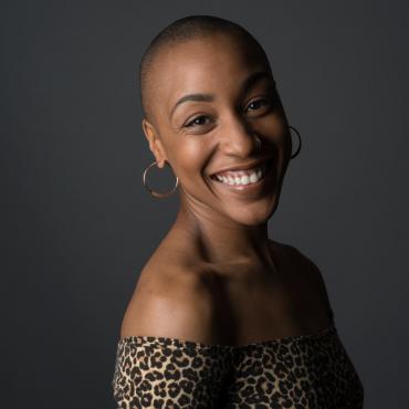 Vie Boheme, a thirty-something Black woman, multimodal performance artist with a bald fade hair cut smiling for a headshot over her right shoulder.